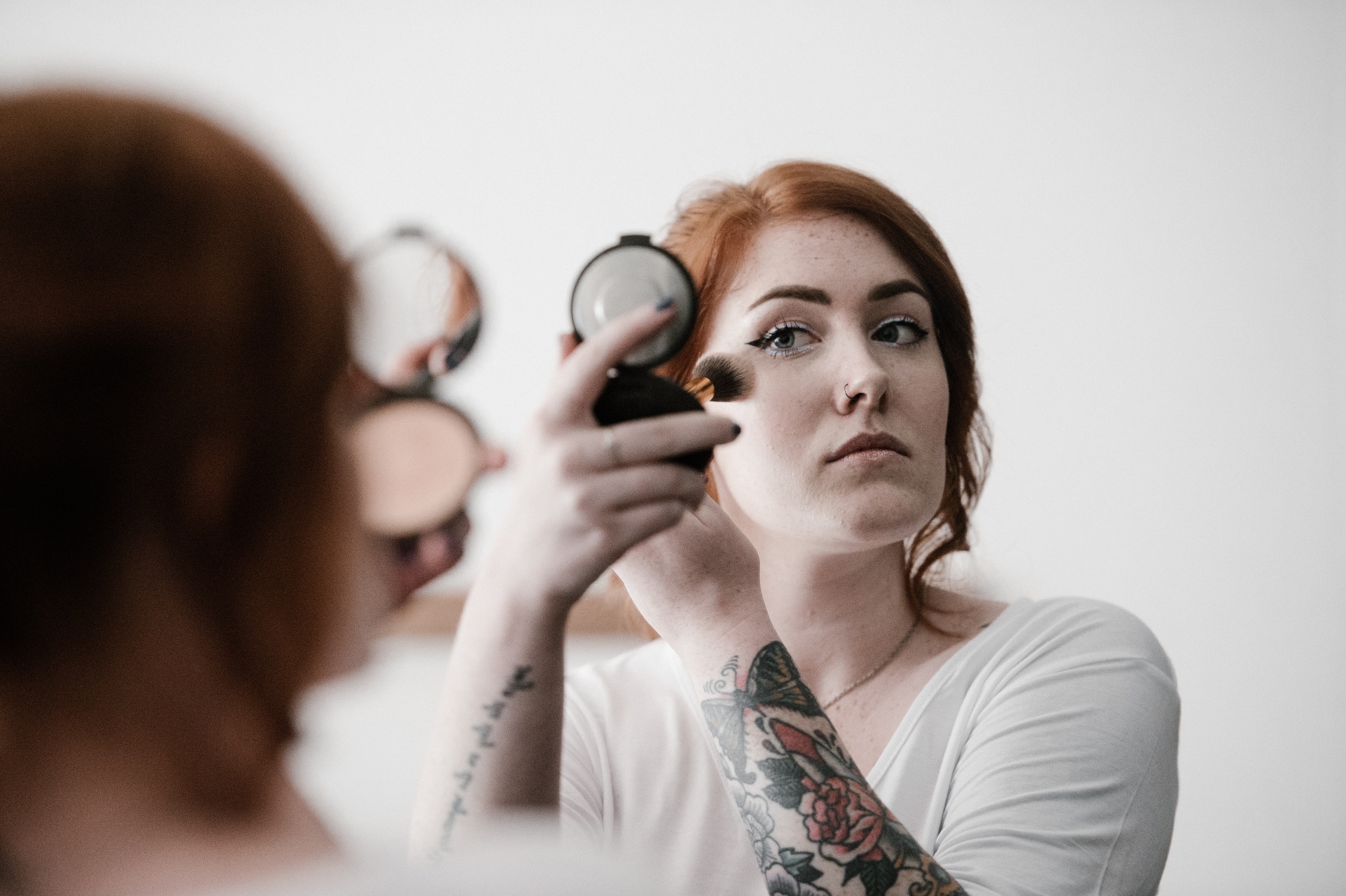 O femeie cu tatuaje se machiaza cu o pensula in timp ce se uita in oglinda.